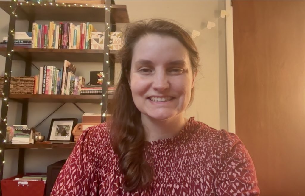 Beth sits smiling in a red and white patterned shirt in front of a bookshelf decorated with books, small boxes, and string lights. A paper heart chain links the bookshelf and a brown door that is partially in view.